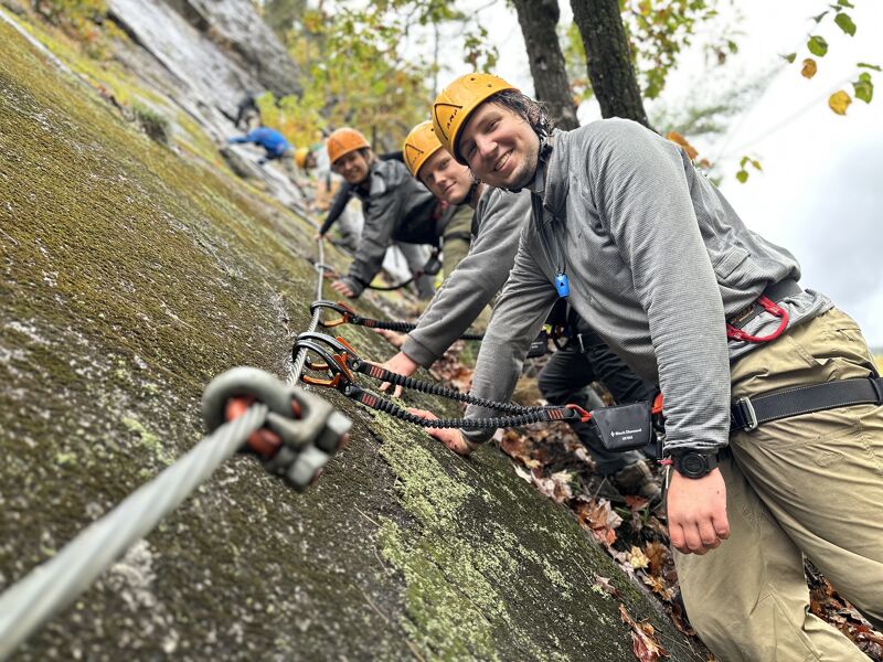 The image shows three people wearing helmets and climbing on a rocky cliff face. They are attached to a safety cable with carabiners. The person in the foreground is smiling and looking at the camera. The background includes trees and a cloudy sky. It appears they are enjoying a Via Ferrata activity.